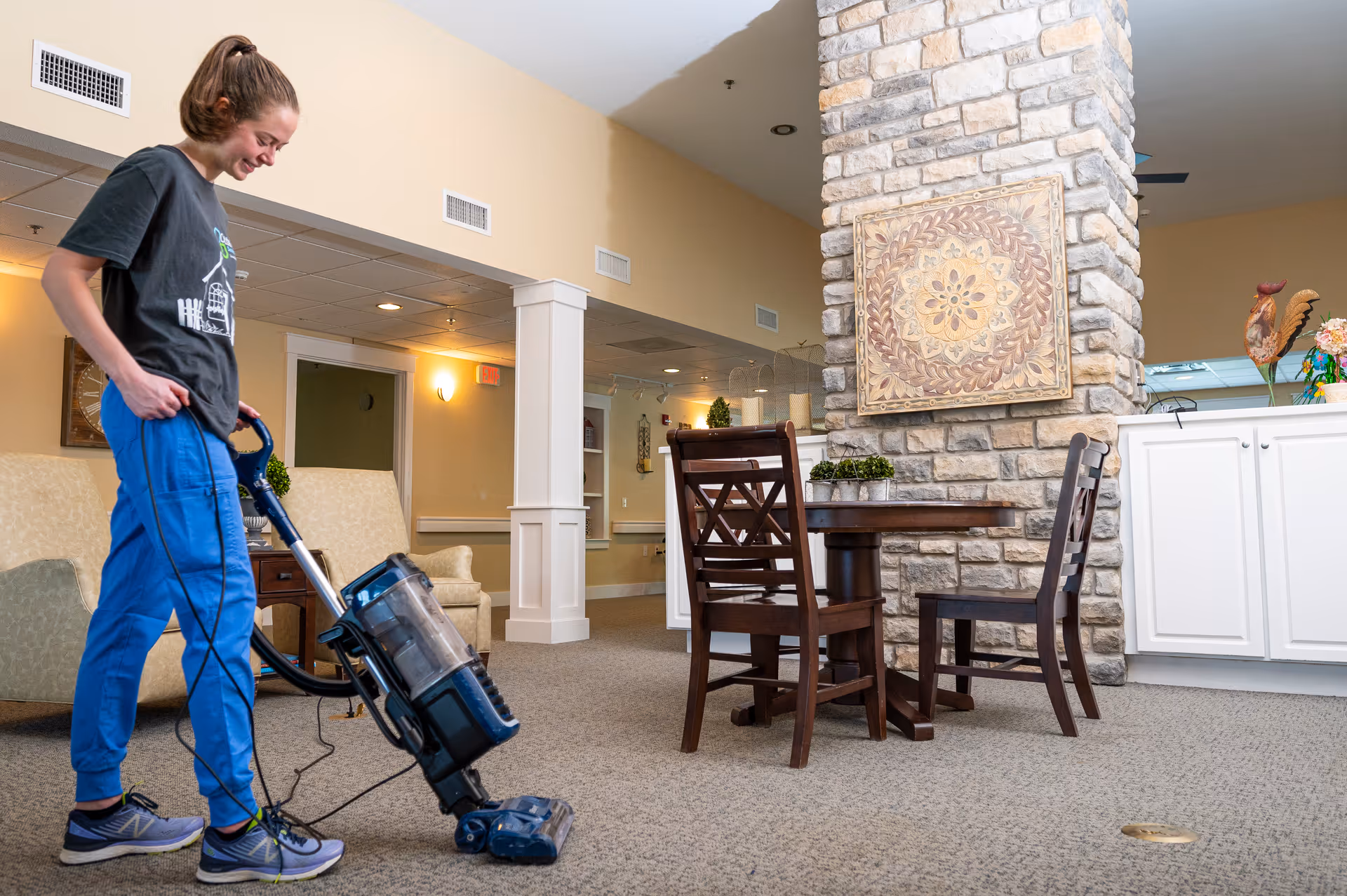 A young woman vacuuming the carpet in a senior living facility common area. The room features a stone pillar with decorative artwork, a wooden table with chairs, beige walls, and comfortable seating. The woman is wearing a dark t-shirt and blue pants, and she is smiling while cleaning.