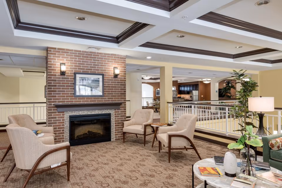 A cozy seating area in a senior living facility featuring four beige armchairs arranged around a brick fireplace with a framed picture above it. The room has a patterned carpet, recessed ceiling lights with dark wood trim, and a railing overlooking a lower level. A small round table with books and a lamp is visible in the foreground, along with some green plants.