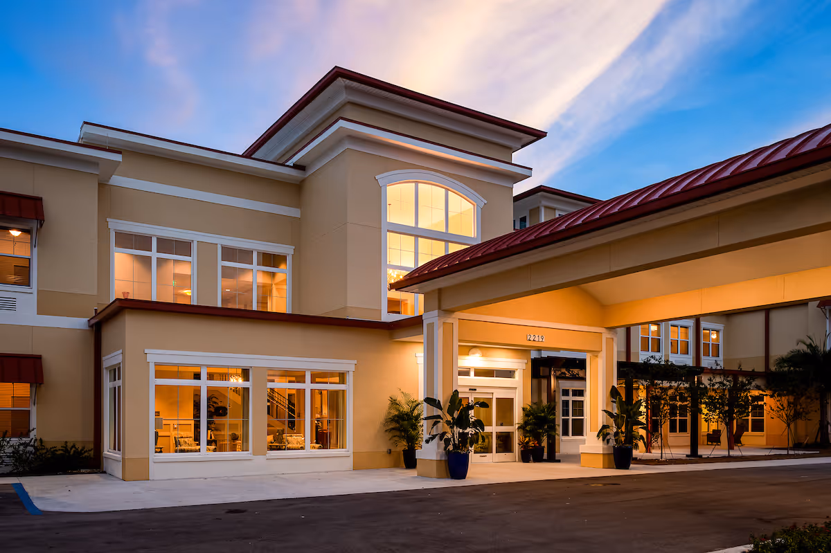 Exterior view of The Gallery at Cape Coral senior living facility at dusk, showing a well-lit entrance with large windows, beige walls, red roof accents, and potted plants near the entrance.
