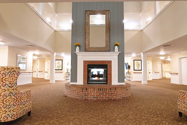 Spacious senior living facility common area with a central brick fireplace, a large mirror above it, and two floral armchairs on either side. The area has beige walls, patterned carpet, and is well-lit with ceiling lights. Hallways and doors are visible in the background.