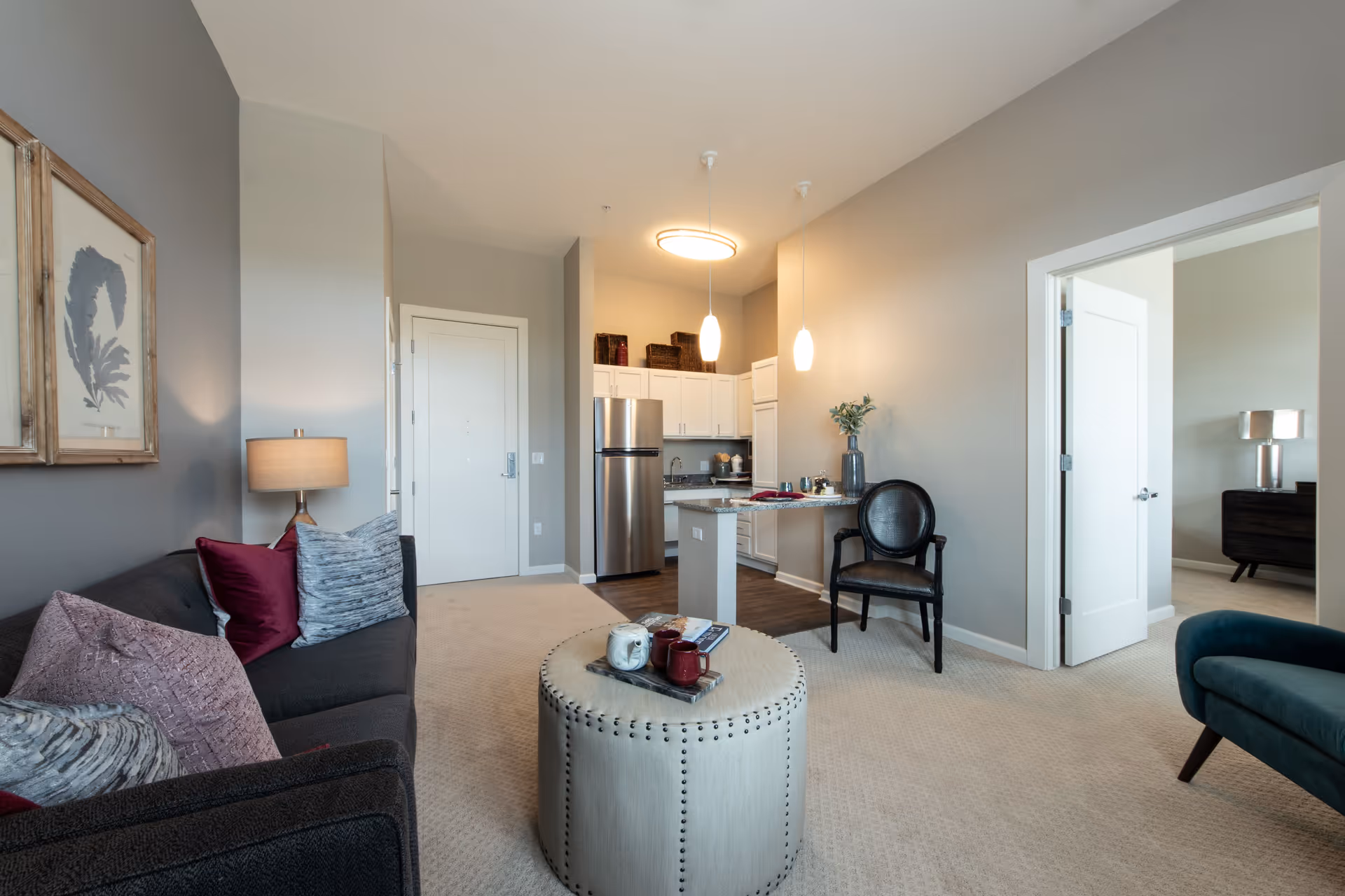 A modern living room with a dark gray sofa adorned with various pillows, a round beige ottoman with decorative items on top, a black chair with a vase on a small table beside it, and a view into a kitchen area with white cabinets and stainless steel refrigerator. There is an open doorway leading to a bedroom with a lamp and dresser visible.