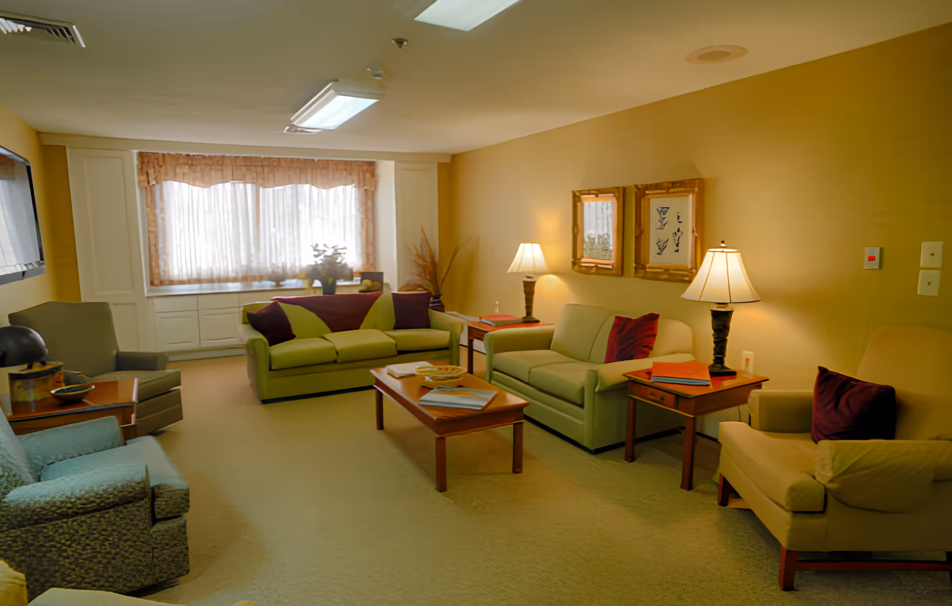 A cozy senior living room with two green sofas, two armchairs, a wooden coffee table with books and a bowl, two side tables with lamps, framed artwork on the wall, and a large window with sheer curtains letting in natural light.