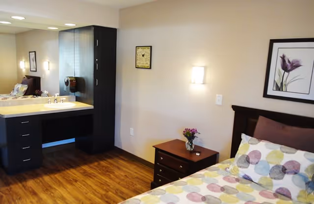A bedroom in a senior living facility featuring a bed with colorful polka dot bedding, a wooden nightstand with a small vase of flowers, a framed floral picture on the wall, and a vanity area with a sink and mirror. The room has wooden flooring and soft lighting.