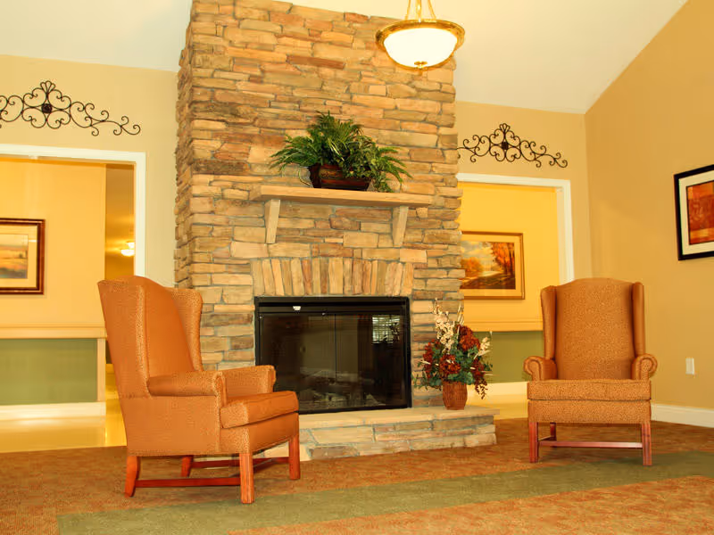 Two upholstered armchairs facing a stone fireplace with decorative plants and wall art in a cozy common room.