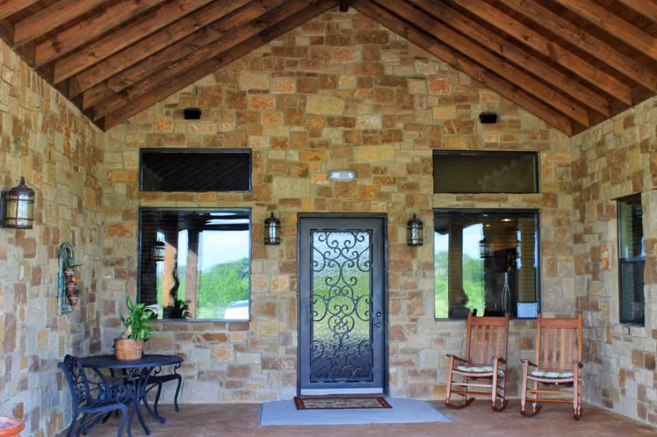Covered outdoor patio area with stone walls and wooden ceiling beams. There is a black metal table with two matching chairs on the left side, a potted plant on the table, and two wooden rocking chairs with cushions on the right side. A decorative black metal door is centered between two windows, each with a small transom window above. Two wall-mounted lantern-style lights flank the door.