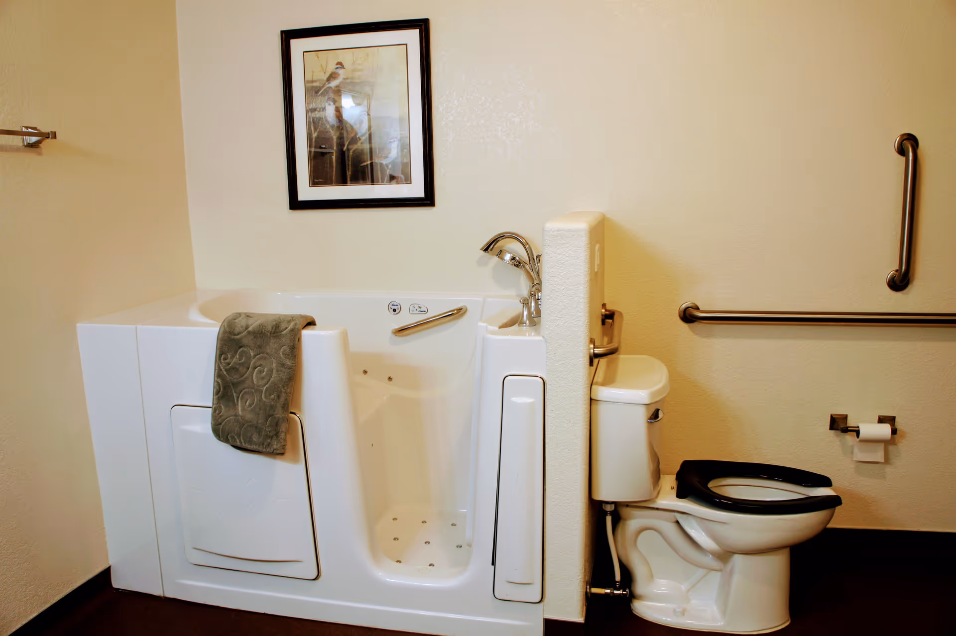 Accessible bathroom featuring a white walk-in bathtub with a towel, an adjacent toilet with a black seat, and wall-mounted grab bars.