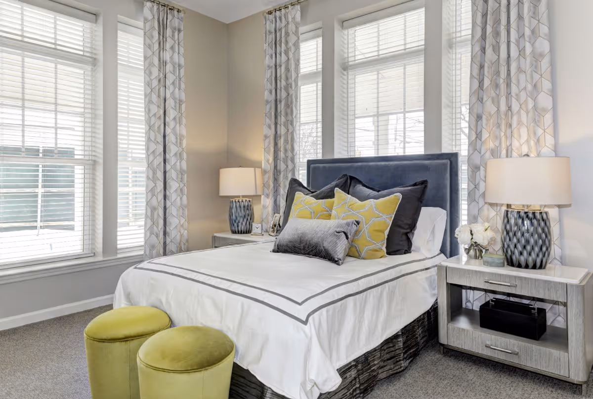 A bright bedroom with a bed featuring a blue upholstered headboard, white bedding with gray trim, and multiple decorative pillows in gray and yellow. There are two green ottomans at the foot of the bed. The room has large windows with white blinds and patterned curtains. Two bedside tables each hold a lamp with a blue and white patterned base, and one table has a small vase with white flowers.