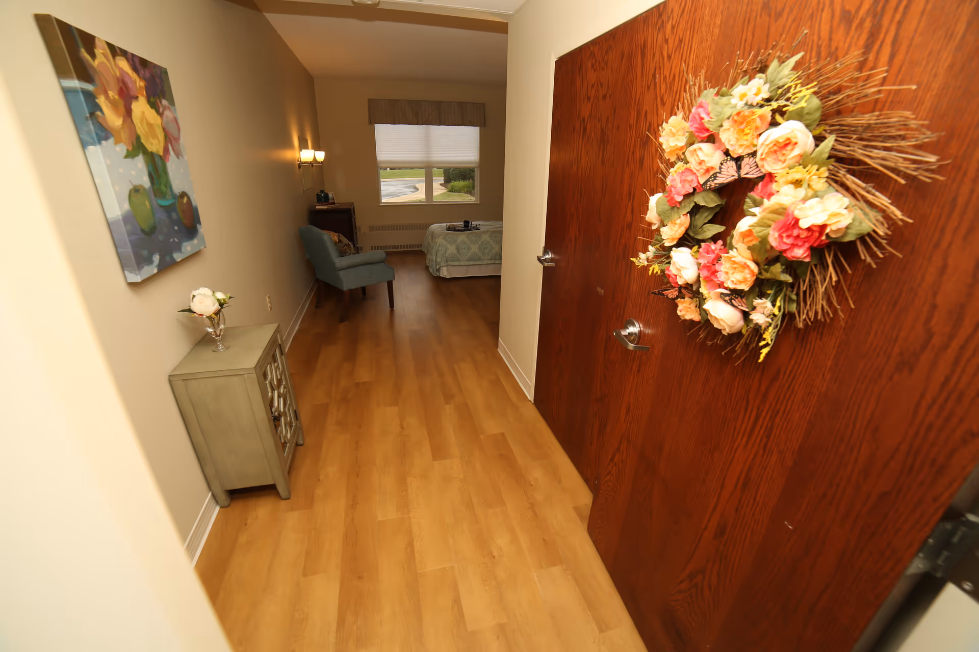 View into a senior living bedroom from the entrance door decorated with a floral wreath. The room has wooden flooring, a blue upholstered chair, a small cabinet with a flower vase, a wall painting of flowers, and a bed with a patterned bedspread near a window with a valance.