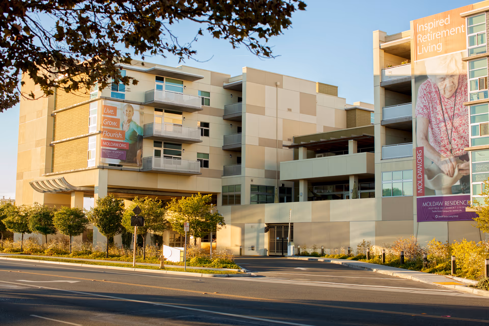 Exterior view of Moldaw Residences, a modern multi-story retirement living facility with large banners displaying elderly individuals and messages promoting inspired retirement living. The building is surrounded by trees and landscaping along a street.