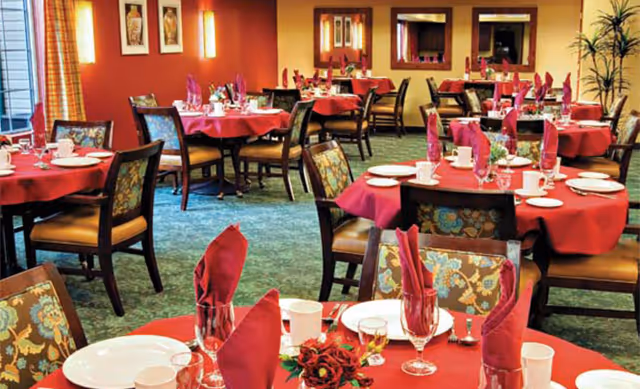 Dining room with multiple round tables covered with red tablecloths, each set with plates, glasses, cups, and red folded napkins. Chairs with floral upholstery surround the tables. The room has warm lighting, framed artwork on the walls, and large mirrors reflecting the space.