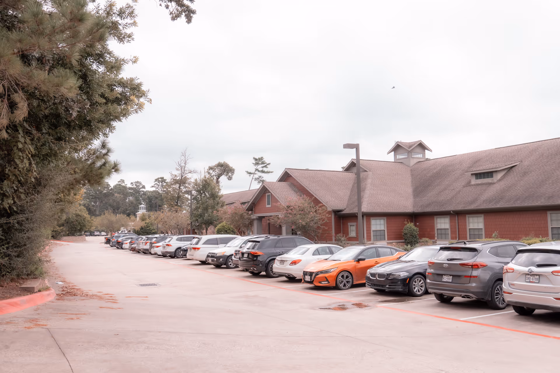 Parking lot filled with cars in front of a single-story red building surrounded by trees.