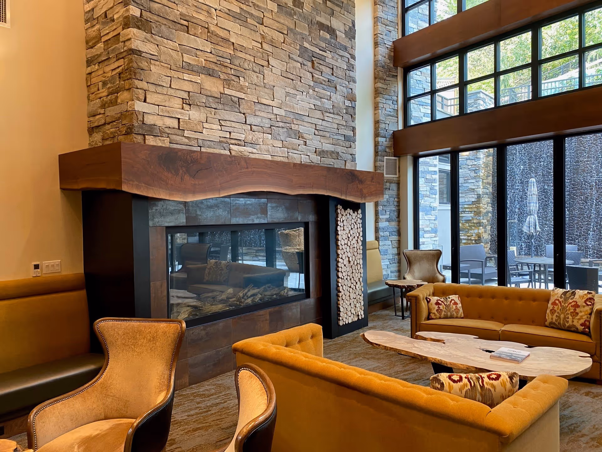 A cozy living room area with mustard yellow sofas and armchairs arranged around a wooden coffee table. There is a large stone fireplace with a wooden mantle, and tall windows letting in natural light and showing an outdoor patio area with tables and chairs.