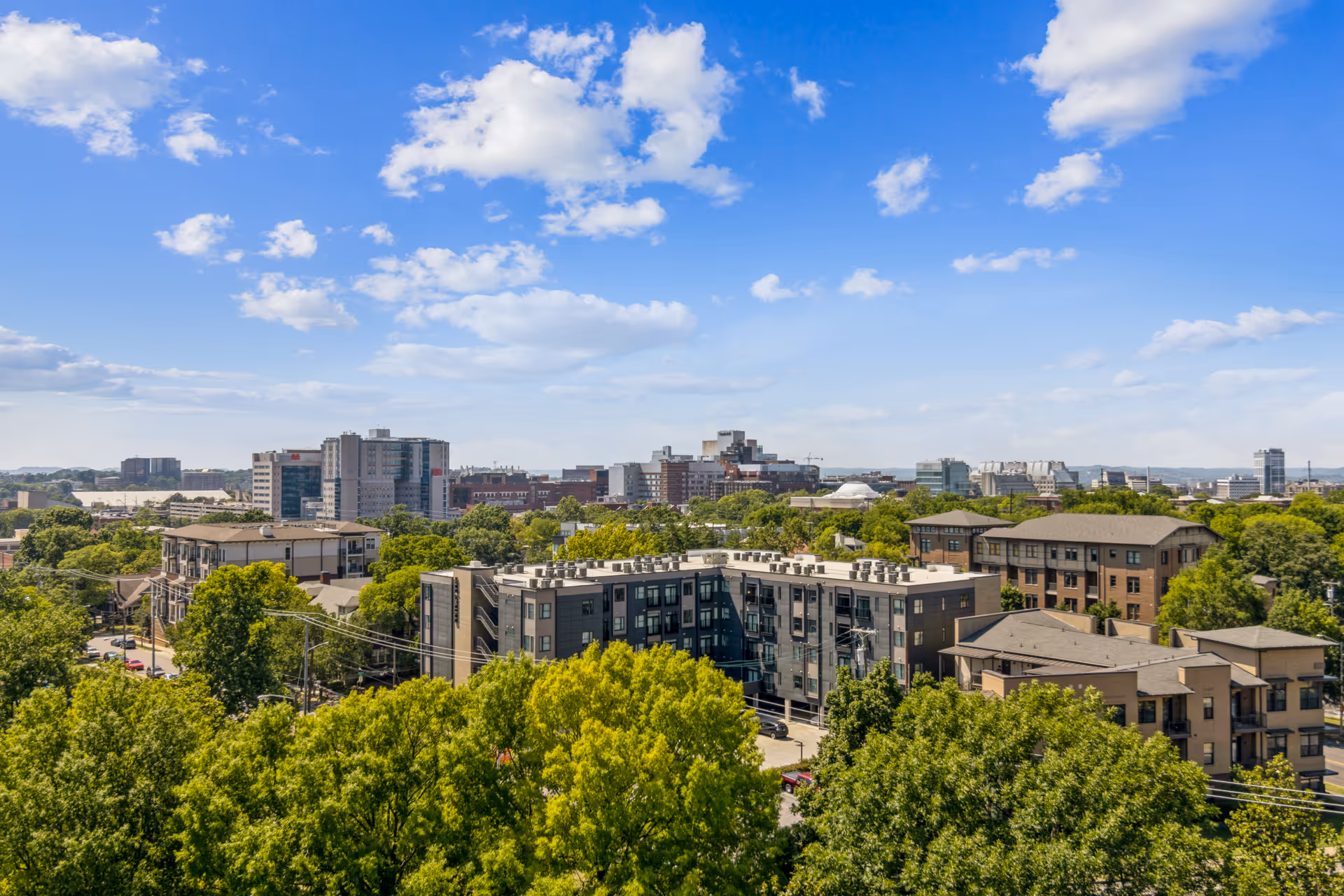 A wide view of a cityscape with multiple buildings surrounded by green trees under a blue sky with scattered clouds.