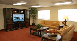 A cozy living room with two beige sofas, a wooden coffee table with a plant centerpiece, a large dark wood entertainment center with a flat-screen TV, a potted plant in the corner, and two windows letting in natural light.