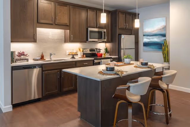 Modern kitchen interior with a central island and bar stools, dark wood cabinets, stainless steel appliances, and pendant lights.