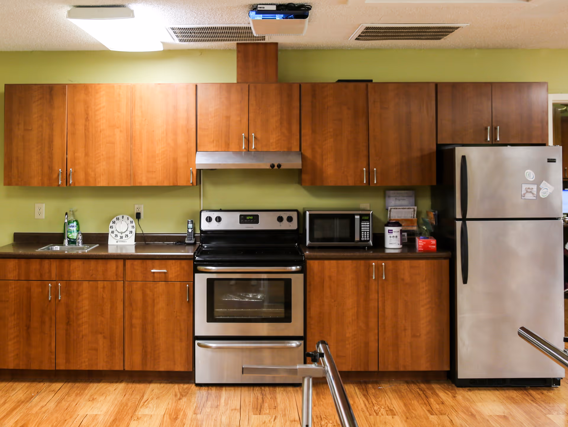 A kitchen area with wooden cabinets, a stainless steel oven and stove, a microwave, a stainless steel refrigerator, a small sink with dish soap, a timer, and a cordless phone on the countertop. The walls are painted light green and the floor is wooden.