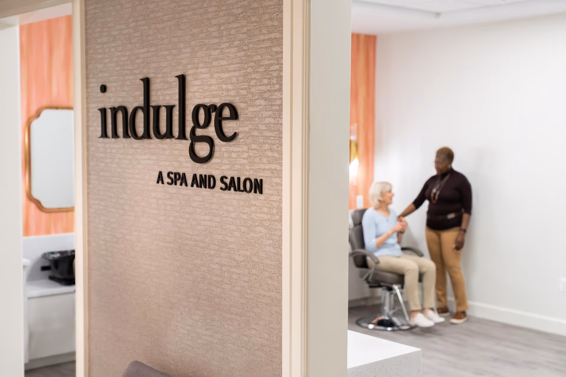 Interior view of a spa and salon area with a wall sign that reads 'indulge A SPA AND SALON'. In the background, a seated elderly woman is holding hands with a standing woman in a welcoming and comforting manner.