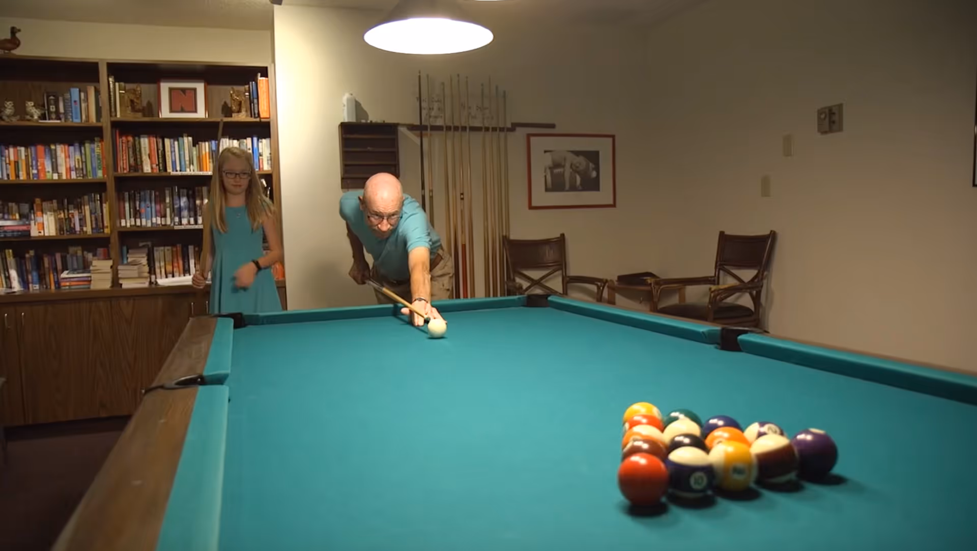An elderly man is playing pool, aiming with a cue stick at the white cue ball on a pool table. A young girl in a blue dress stands nearby watching. Behind them is a bookshelf filled with books and a wall with pool cues and framed pictures. The room is softly lit with a hanging light above the pool table.