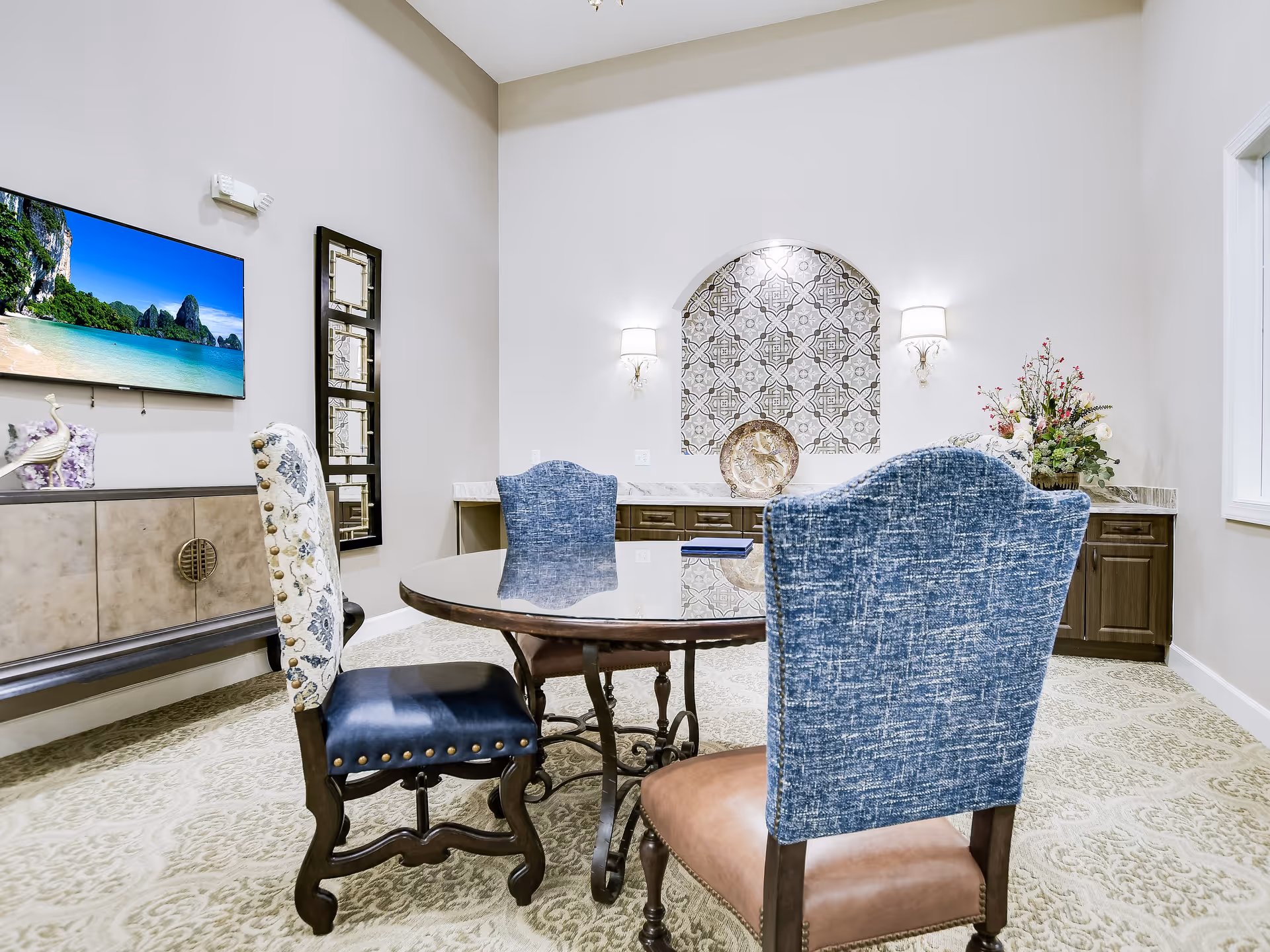 A bright and elegant interior room featuring a round glass-top table with four upholstered chairs in blue and patterned fabric. The room has a decorative tiled alcove on the far wall with two wall sconces, a sideboard with a flat-screen TV mounted above it, and a floral arrangement on a countertop to the right.
