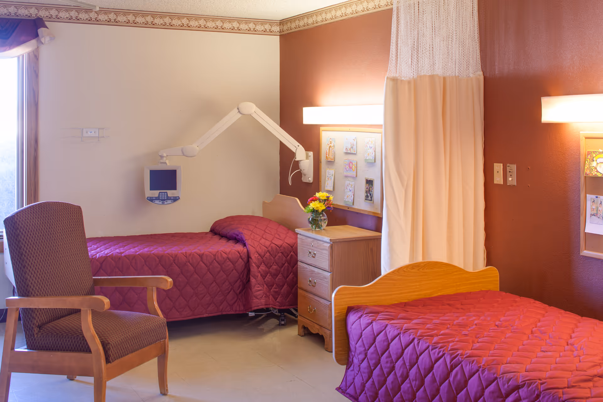 A room with two single beds covered in matching burgundy quilted bedspreads. Between the beds is a wooden nightstand with a vase of flowers on top. A privacy curtain hangs between the beds. There is a padded wooden armchair near a window with a valance. A medical monitor is mounted on the wall above one bed. The walls are painted in beige and burgundy tones, with framed artwork and a bulletin board on the wall.