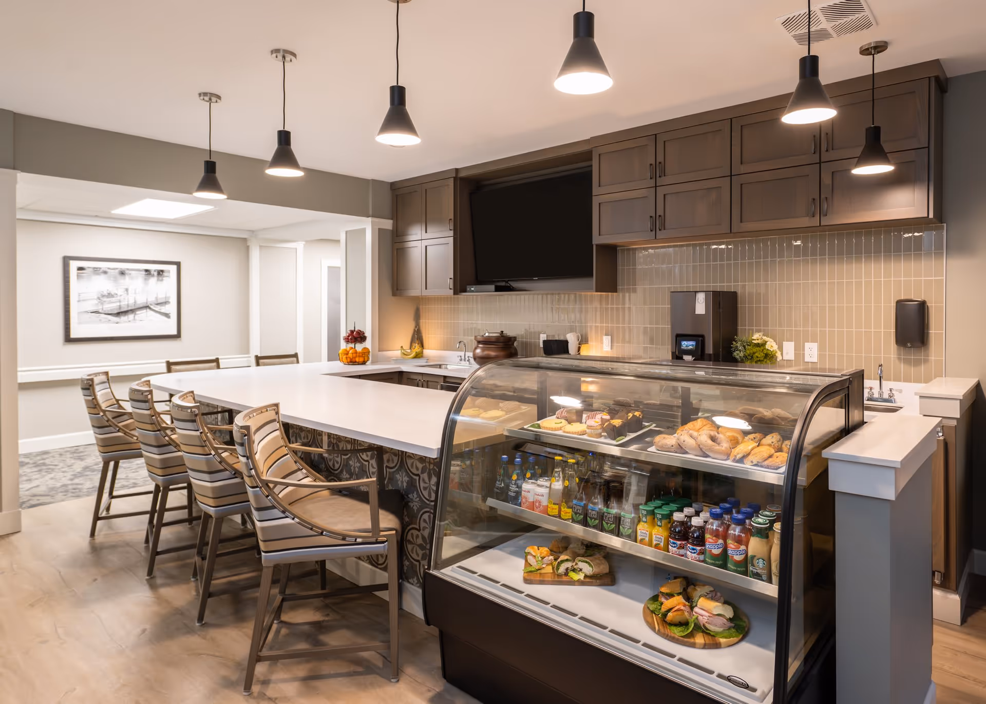Interior view of a modern dining area with a long white countertop surrounded by striped cushioned chairs. A glass display case in the foreground contains various beverages and sandwiches. The background features wooden cabinets, a mounted flat-screen TV, pendant lights hanging from the ceiling, and a framed black and white photo on the wall.