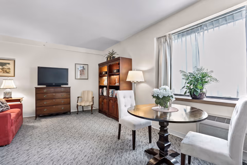 A bright living room with a round wooden table and upholstered chairs by a window, a TV on a dresser, a bookshelf, and a red sofa.