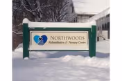 A sign for Northwoods Rehabilitation & Nursing Center mounted on green posts, surrounded by snow with trees and a building in the background.