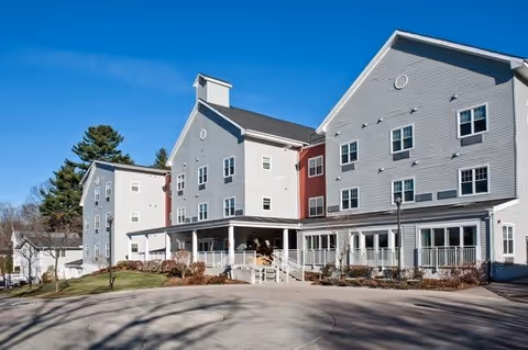 Exterior view of a multi-story senior living facility building with white and gray siding, multiple windows, and a covered entrance. The building is surrounded by a paved driveway and some landscaping with bushes and trees under a clear blue sky.