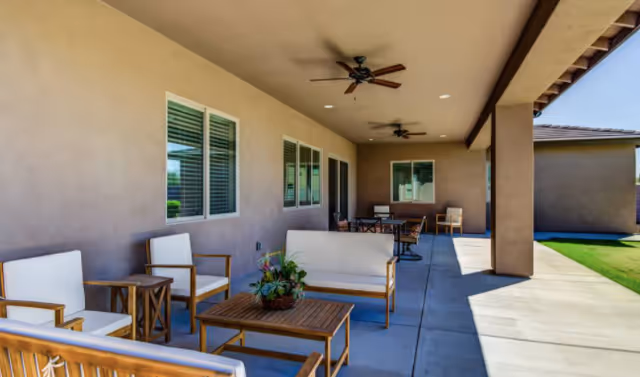 Covered outdoor patio area with ceiling fans, wooden furniture including chairs, a loveseat, and tables, and a potted plant on the table. The patio overlooks a grassy area and is attached to a building with beige walls and windows.