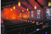 Interior view of a chapel with wooden pews arranged in rows facing an altar. The ceiling has exposed wooden beams and hanging pendant lights. Stained glass windows are visible on the right side, allowing colorful light to enter the space.