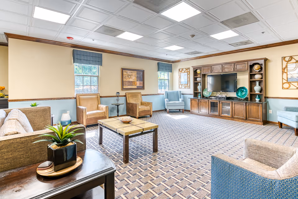Bright senior living common lounge with sofas, armchairs, a central coffee table, and an entertainment cabinet with TV.