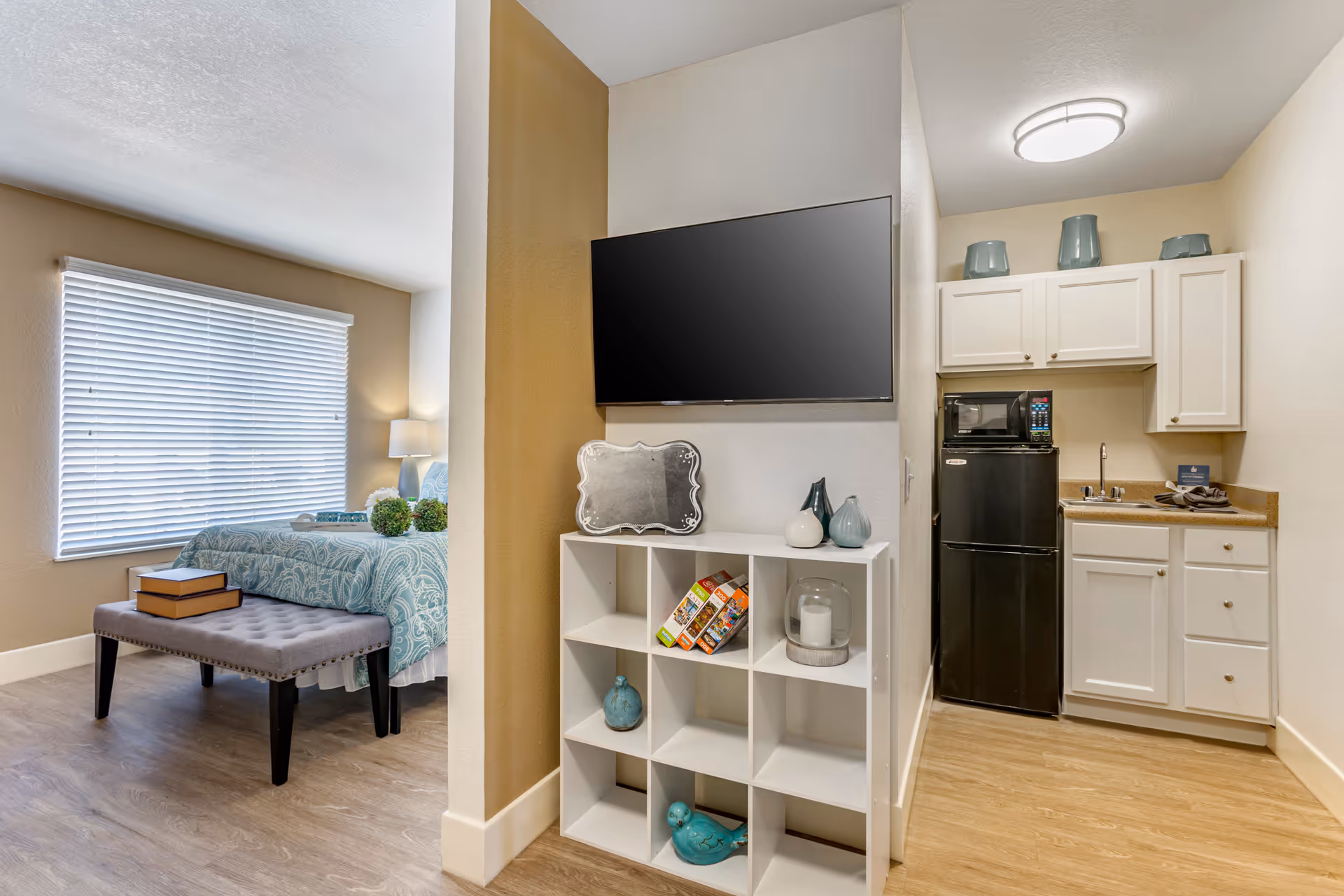 A cozy studio apartment with a small kitchenette featuring a black refrigerator, microwave, and white cabinets. Adjacent to the kitchenette is a white cubby shelf with decorative items and a mounted flat-screen TV above it. To the left, there is a sleeping area with a bed covered in a blue patterned comforter, a gray bench at the foot of the bed with two books on it, and a window with closed blinds letting in natural light.