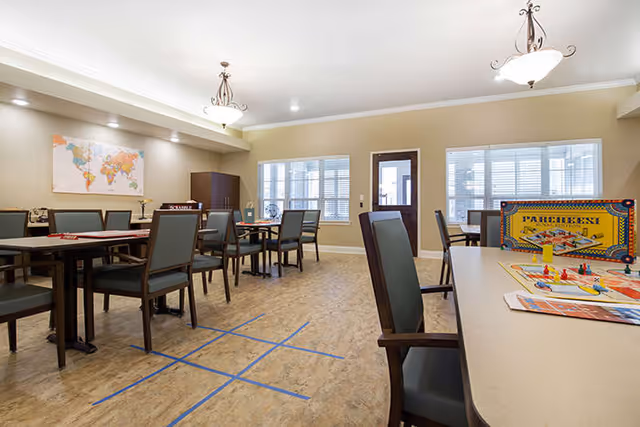 A bright and spacious common room with multiple tables and chairs arranged for group activities. A board game is set up on one table in the foreground, and a world map hangs on the wall. Large windows allow natural light to fill the room.