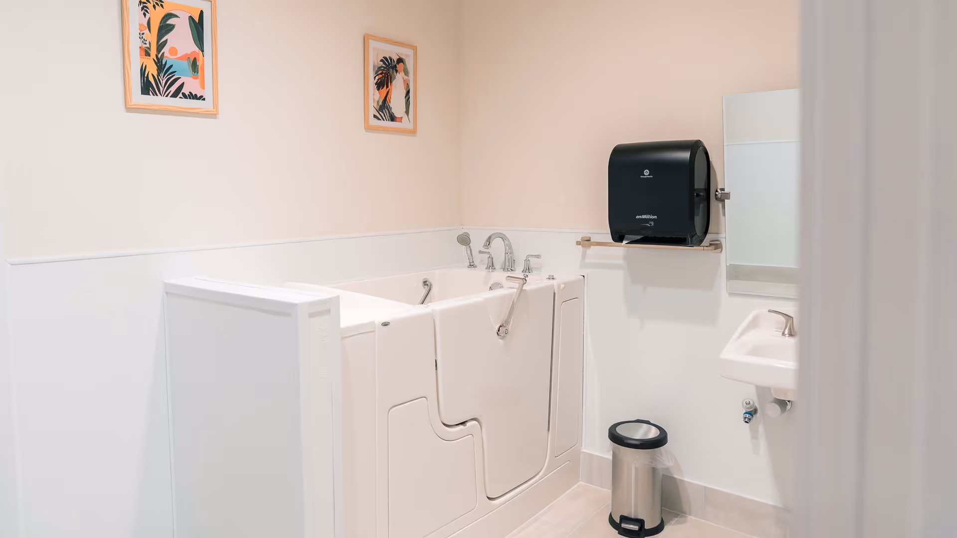 A clean, accessible bathroom featuring a walk-in bathtub with safety handles, a small sink with a faucet, a wall-mounted paper towel dispenser, a trash can, and two framed botanical prints on the wall.