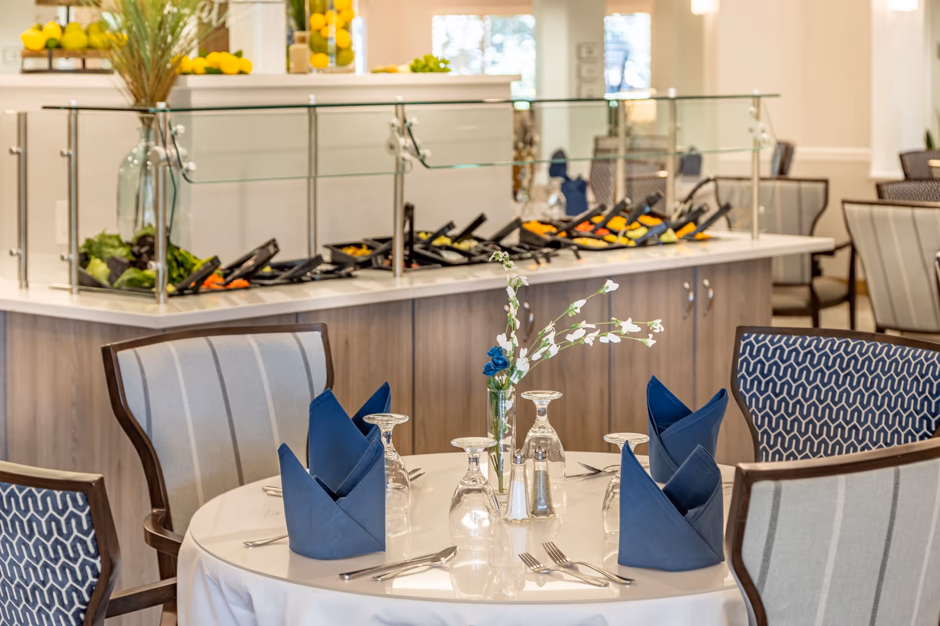 A dining area in a retirement residence featuring a round table set with blue folded napkins, upside-down wine glasses, silverware, and a small vase with white and blue flowers. In the background, there is a buffet counter with a glass sneeze guard displaying various food items and decorative elements like lemons and plants.