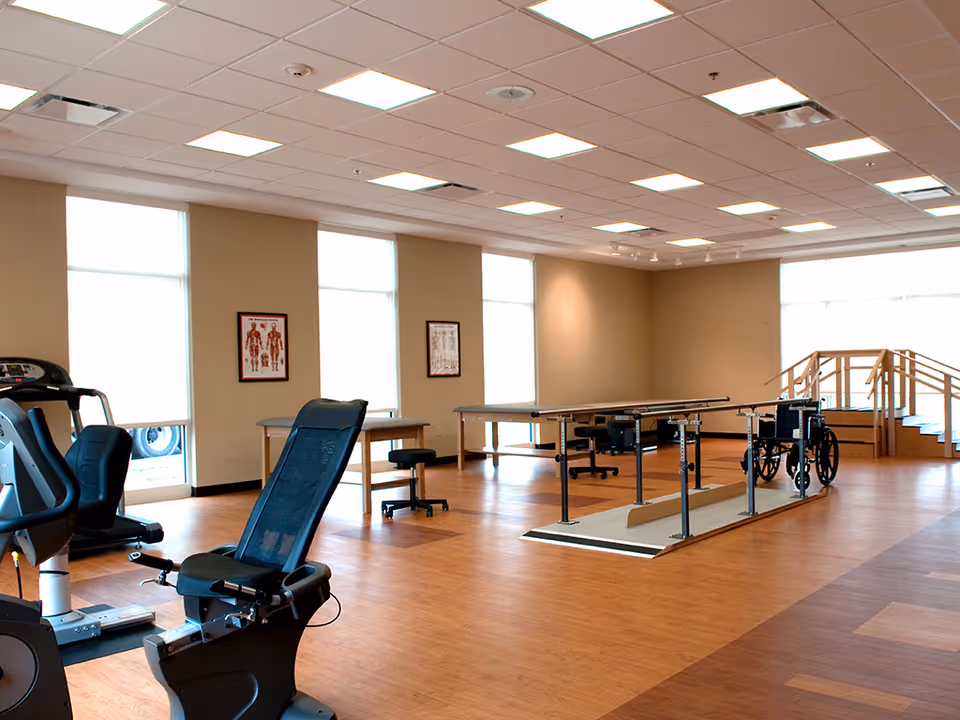 A spacious rehabilitation room with exercise equipment including a recumbent bike, parallel bars for walking practice, a wheelchair, and therapy tables. The room has large windows with natural light, beige walls, and wooden flooring.