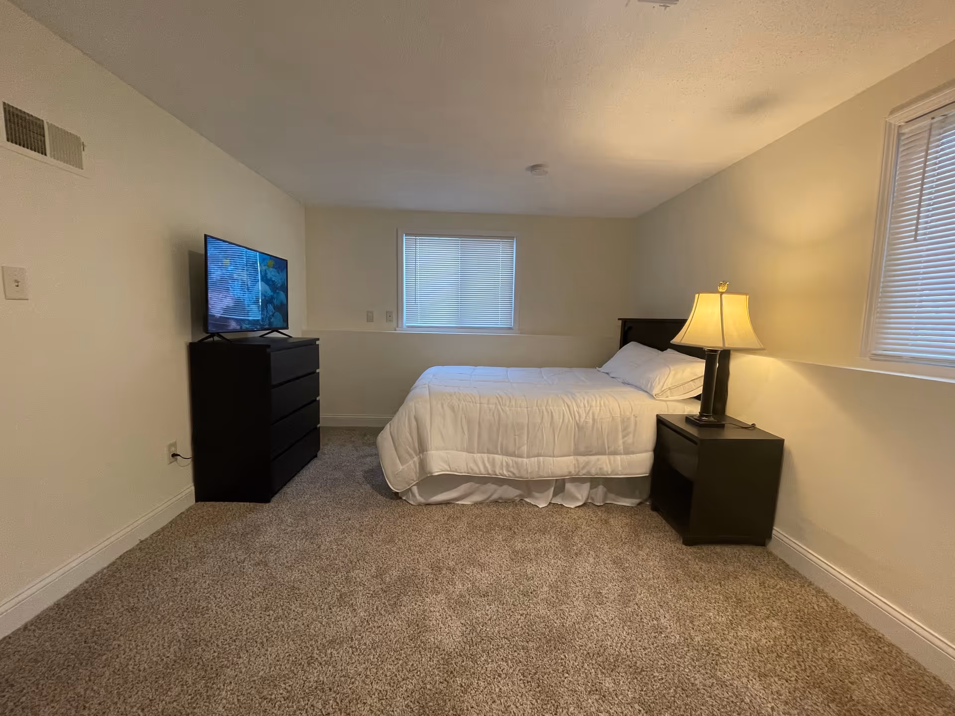 A simple bedroom with beige walls and carpeted floor. The room contains a bed with white bedding positioned against the far wall, a black nightstand with a lamp on the right side of the bed, and a black dresser with a flat-screen TV on top on the left side of the room. Two windows with blinds are visible, one behind the bed and one on the right wall.