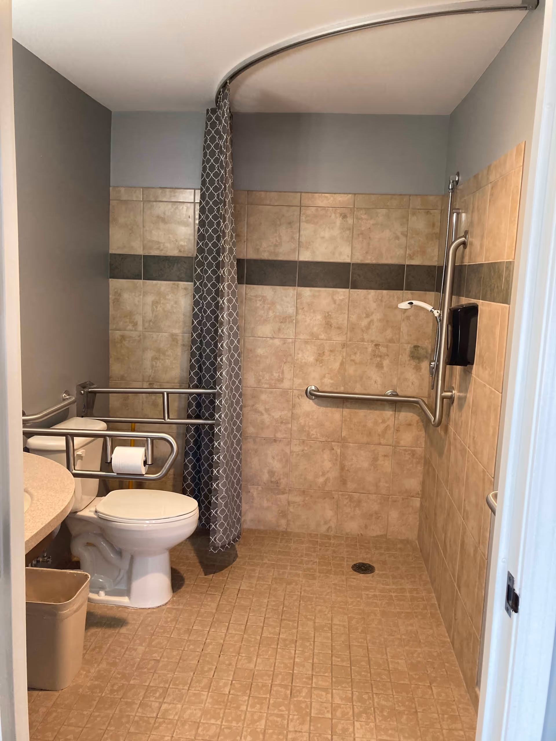 Accessible bathroom with beige tiled floor and walls, featuring a toilet with safety grab bars, a shower area with a handheld showerhead and a patterned shower curtain on a curved rod, a countertop with a sink, and a small trash bin.