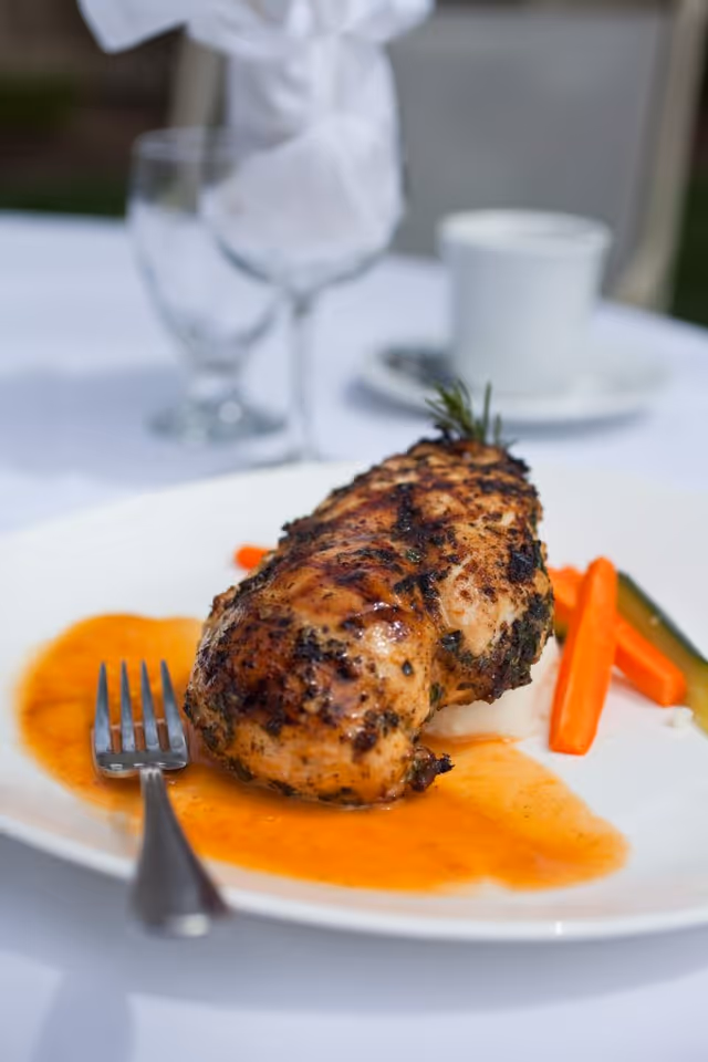 A plated meal featuring a grilled chicken breast with herbs, served with a side of cooked carrots and zucchini, and a sauce on the plate. In the background, there is a glass of water, a wine glass with a folded white napkin, and a white cup on a saucer, all set on a white tablecloth.