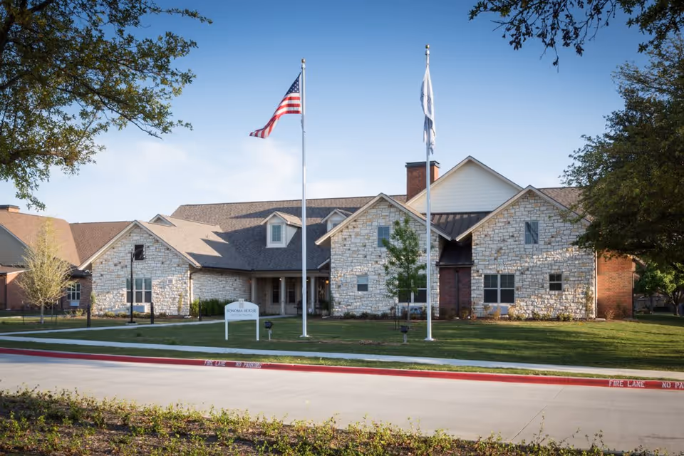 Exterior view of Sonoma House Assisted Living Alzheimer’s Care facility, showing a large stone and brick building with a pitched roof, two flagpoles with an American flag and another flag, surrounded by green lawn and trees under a clear blue sky.