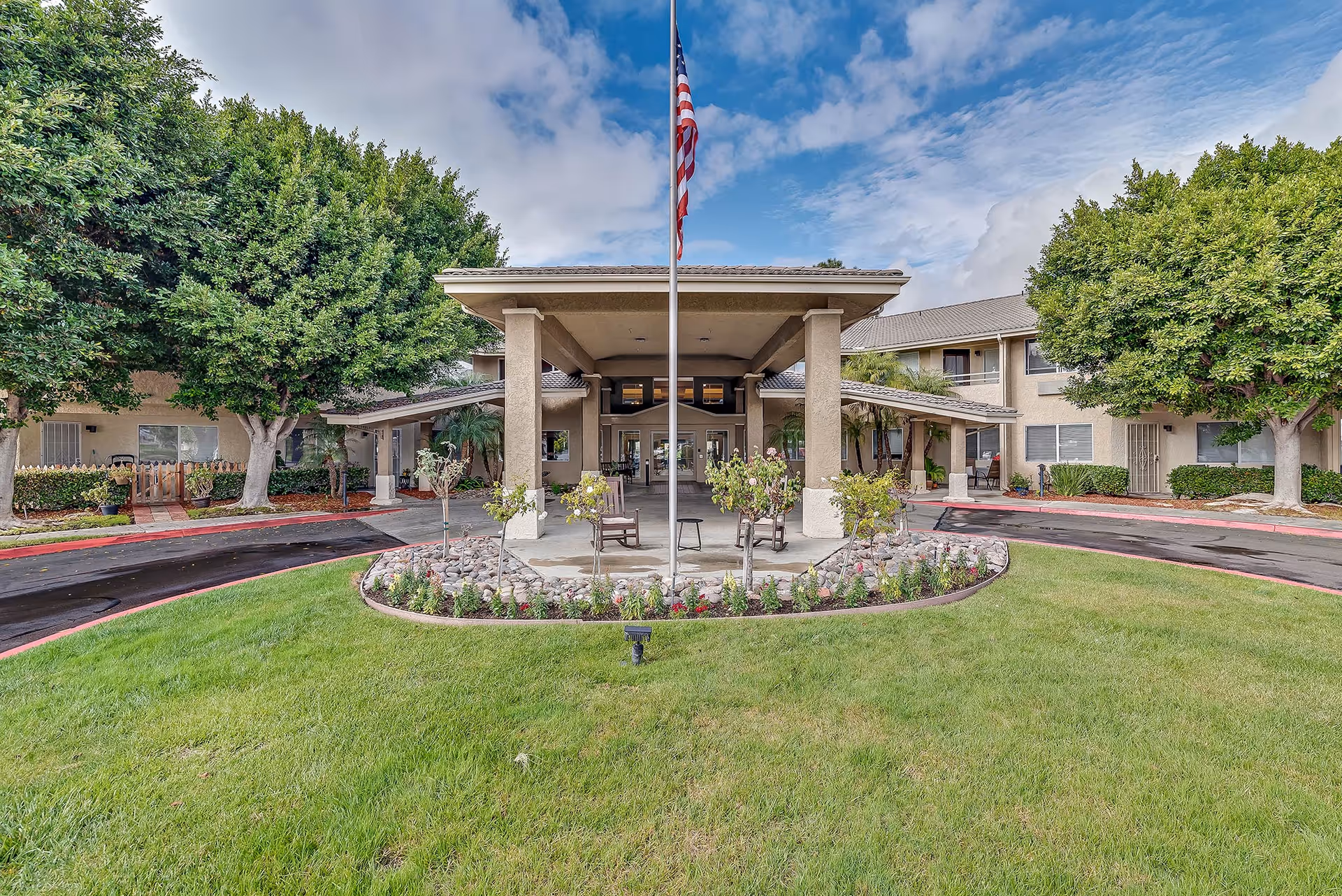 Front entrance of a senior living building with a covered porte-cochère, central flagpole, circular driveway and landscaped lawn.