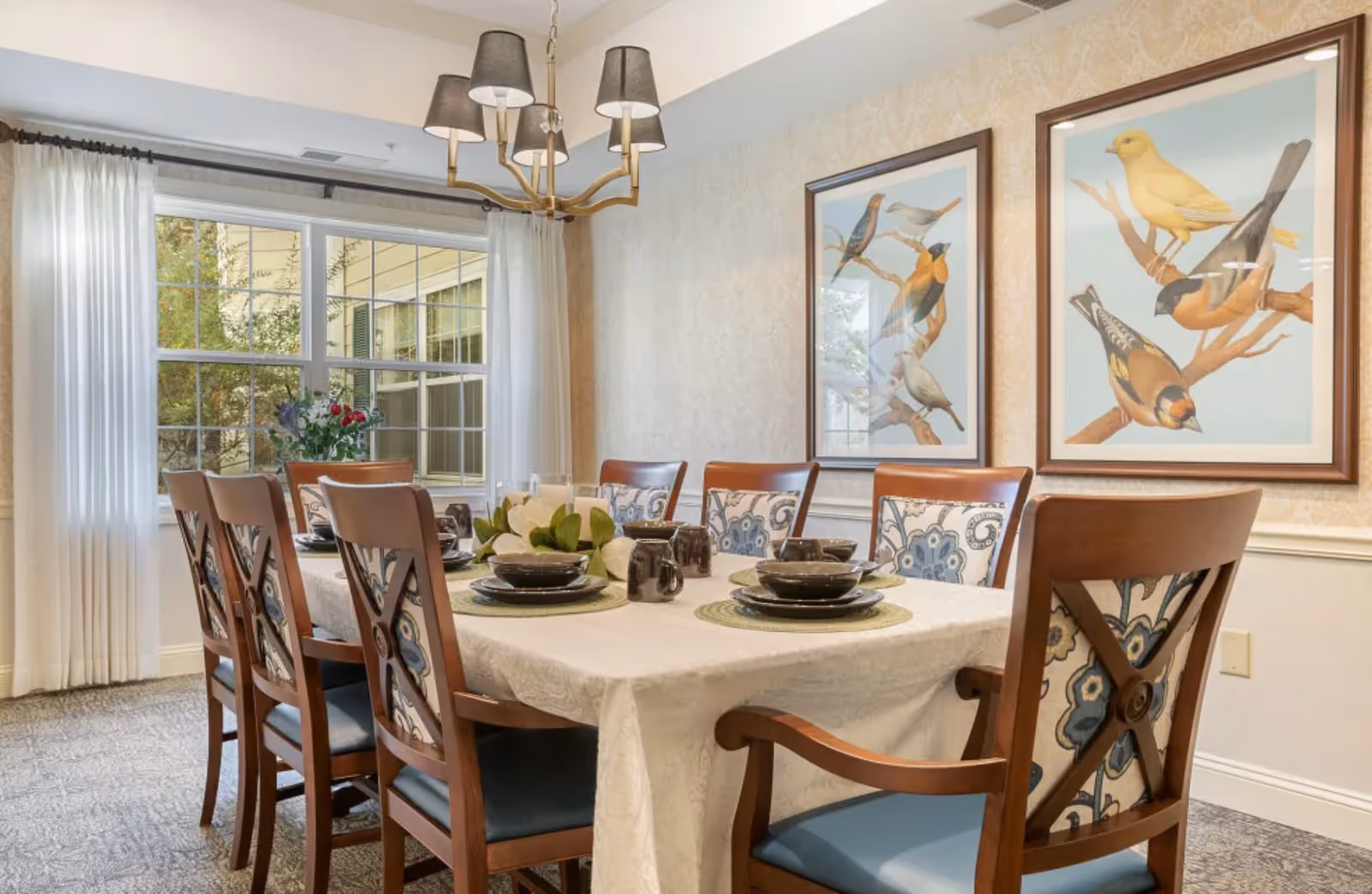 A dining room with a rectangular table covered with a white tablecloth set for six people. The chairs have wooden frames with blue and floral patterned cushions. On the table are black dishes, mugs, and a centerpiece with white flowers. The room has a large window with white curtains and two framed pictures of birds on the wall. A chandelier with five lampshades hangs above the table.
