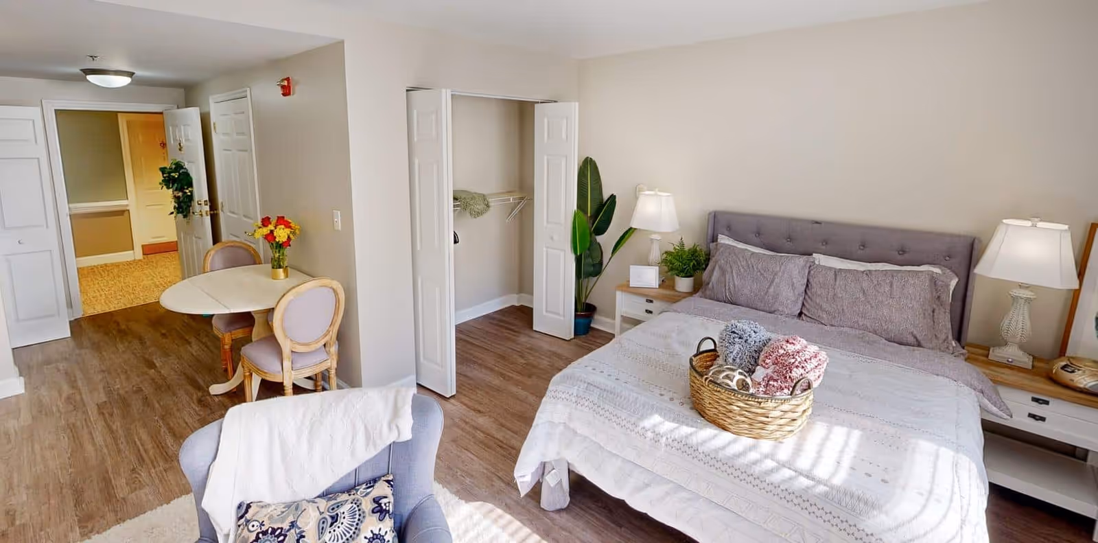 A bright and cozy bedroom in a senior living facility with a neatly made bed featuring gray pillows and a white patterned bedspread. A basket with folded blankets sits on the bed. There are two bedside tables with lamps and a potted plant. An open closet with white double doors is visible, along with a small round dining table with two chairs and a vase of flowers. The room has wood flooring and light-colored walls.
