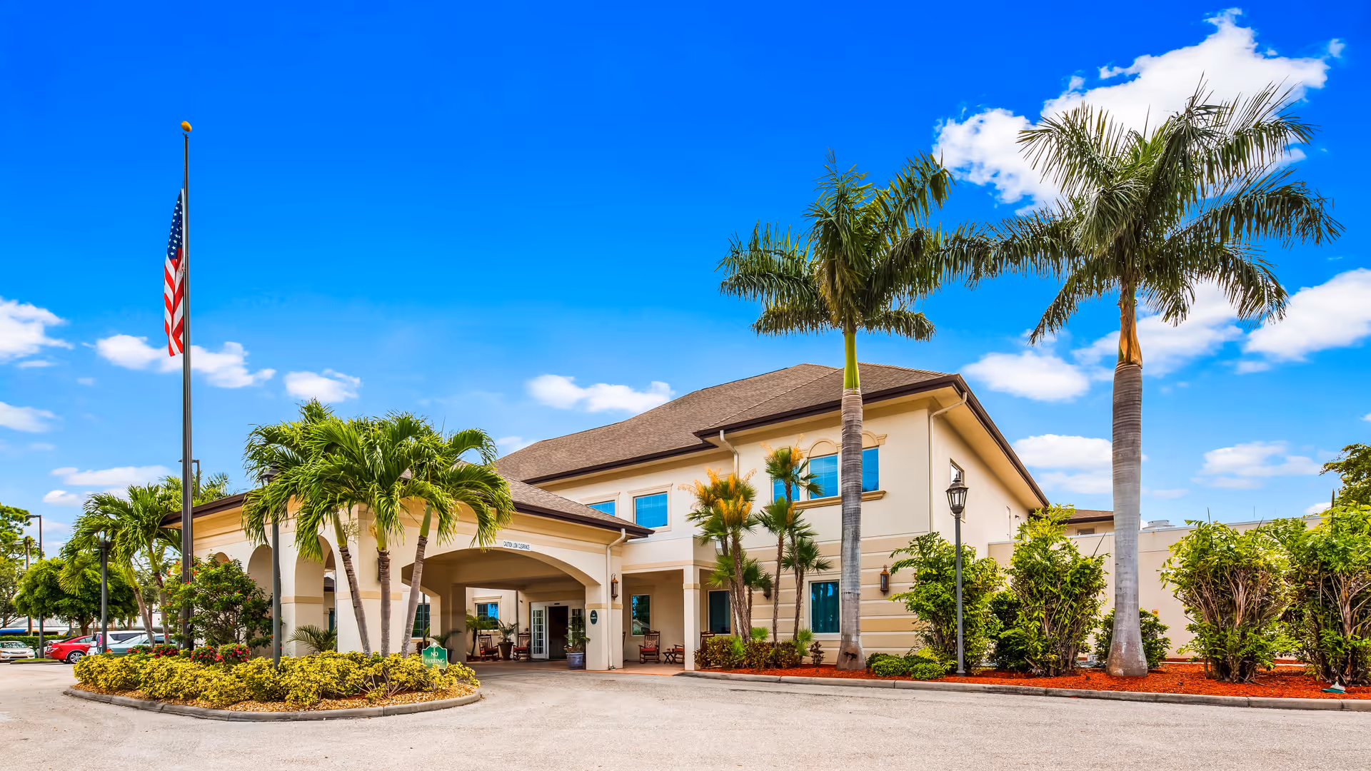 Exterior view of The Windsor of Cape Coral senior living facility with a clear blue sky, palm trees, an American flag on a flagpole, and a driveway leading to the entrance under a covered portico.