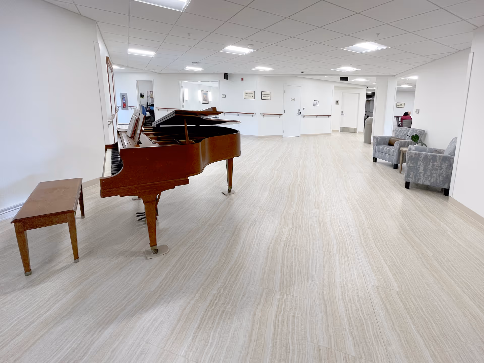 A spacious, well-lit interior area with a grand piano and matching bench on the left side. The floor has a light wood or stone pattern, and the walls are white. On the right side, there are two gray patterned armchairs with a small table between them. The ceiling has recessed lighting and a drop ceiling design. In the background, there are several doors and a person sitting in a chair near the far right wall.