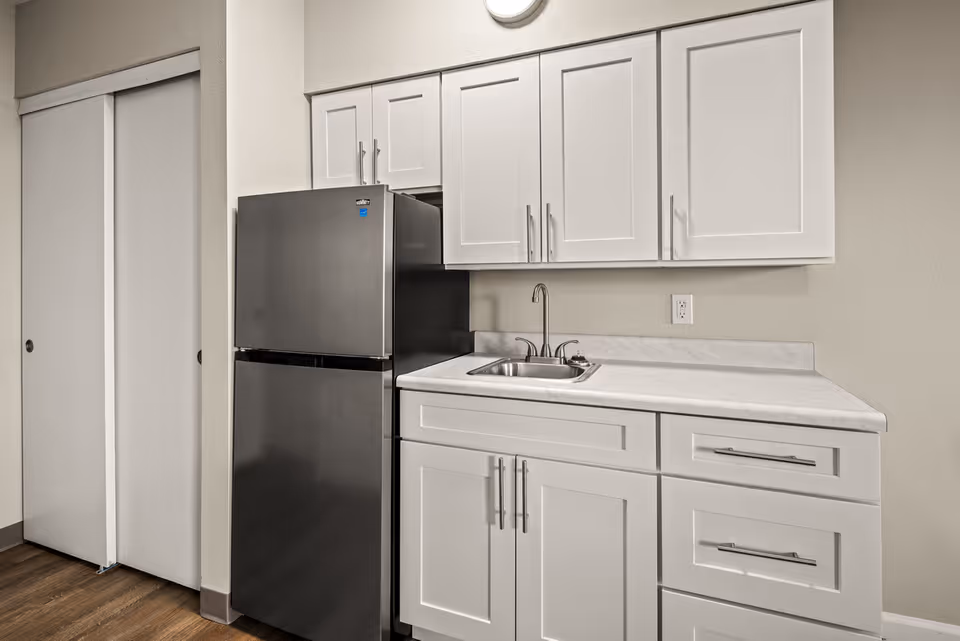 A compact kitchen area with white cabinets, a stainless steel sink, and a black refrigerator. The countertop is light-colored, and there are sliding closet doors to the left. The walls are painted beige and the floor has a wood finish.