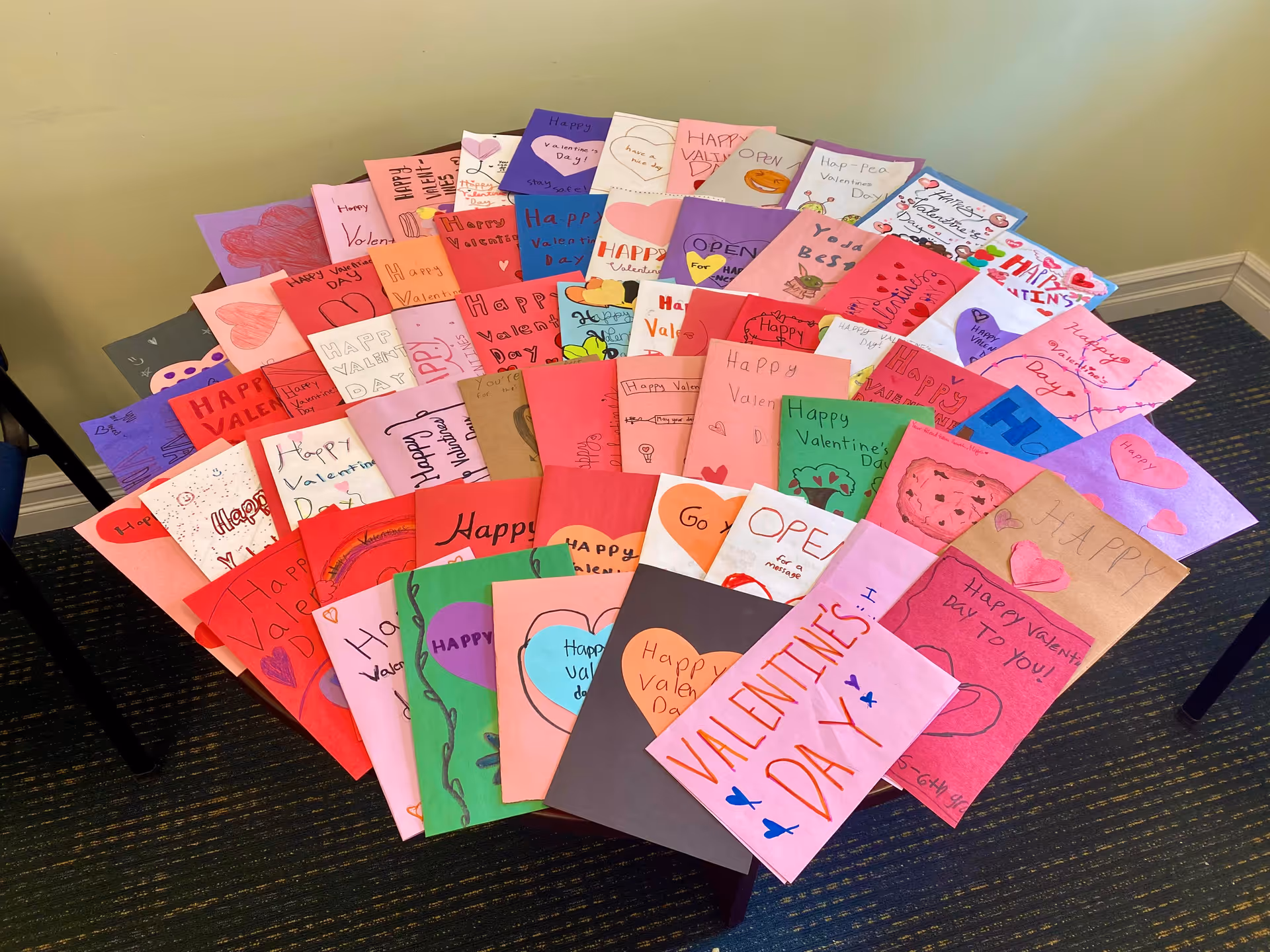A table covered with numerous colorful handmade Valentine's Day cards arranged in rows. The cards feature various designs, hearts, and handwritten messages wishing Happy Valentine's Day. The setting appears to be indoors with a carpeted floor and beige walls.