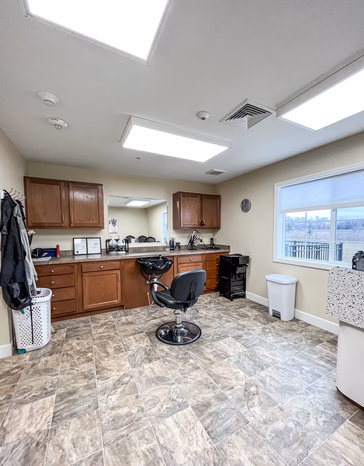 Interior of a hair salon room in an assisted living facility with a single black salon chair in front of a counter with a sink, wooden cabinets above and below the counter, a large mirror on the wall, a window with a view outside, and tiled flooring.