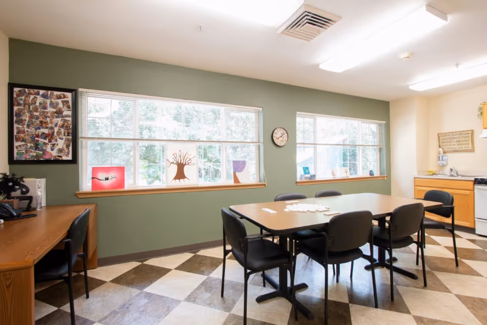 A bright room with large windows letting in natural light, featuring a rectangular table surrounded by six black chairs. The room has a green accent wall with framed artwork and decorative pieces on the windowsill. There is a wooden desk with a chair on the left side and a small kitchen area with a stove and cabinets on the right.
