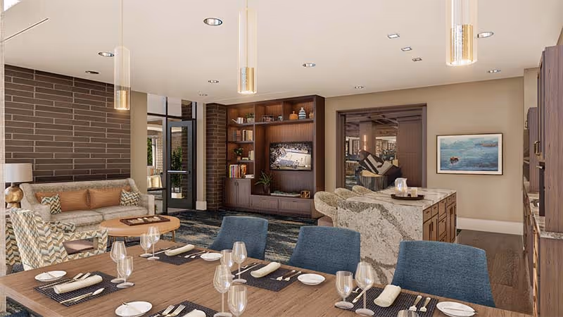 Open-plan communal living area with a set dining table in the foreground, sofas, built-in TV shelving and a kitchen island in the background.